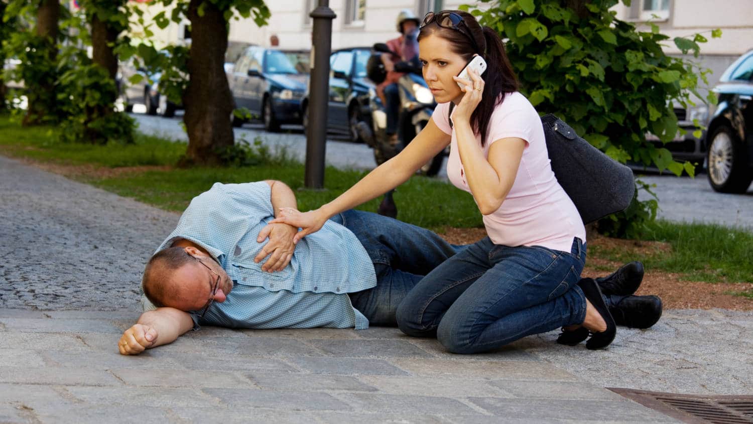 Na zdjęciu widać leżącego mężczyznę trzymającego się za serce. Obok niego klęczy kobieta trzymająca telefon przy uchu. Dzwoni ona po pogotowie 